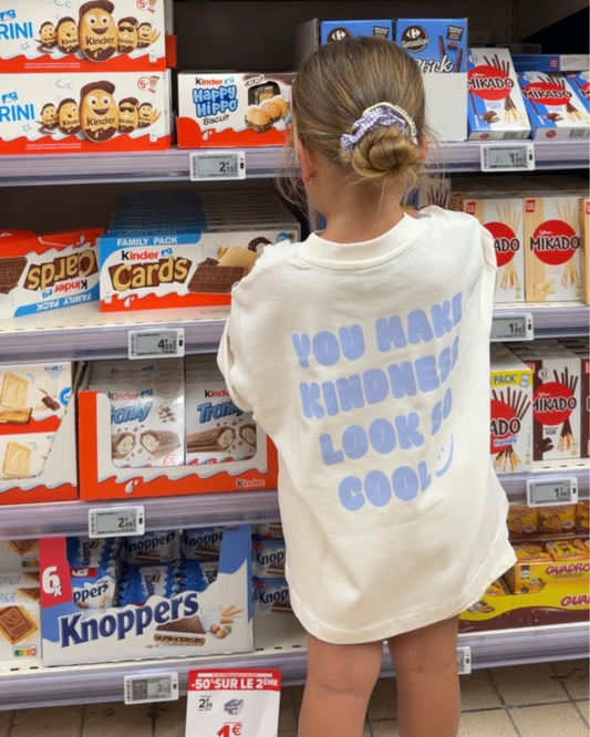 Weißes Oversize-Kindershirt mit blauem Statement-Print „You Make Kindness Look So Cool" – Kind von hinten vor Kinder- und Oreo-Süßigkeitenregal im Supermarkt