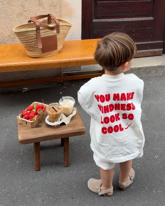 Weißes Oversize-Kindershirt mit rotem Statement-Print „You Make Kindness Look So Cool" – Kind von hinten vor Holztür, sommerliches Café-Setting mit Erdbeeren und Iced Coffee
