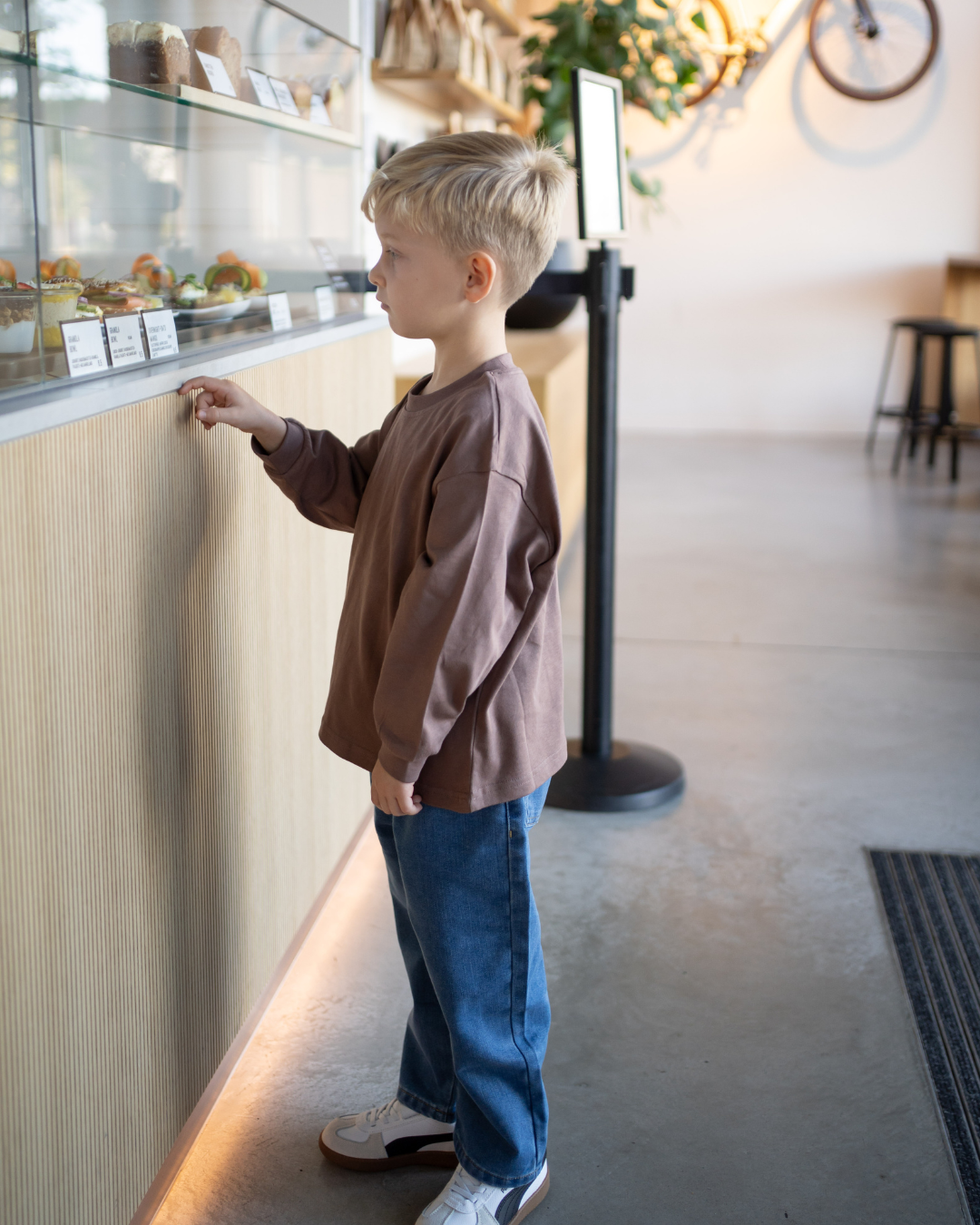 Junge steht seitlich an einer Café-Theke im kompletten Espresso-Look – espressobraunes Longsleeve und blaue Oversize-Jeans von Comfy Studios.