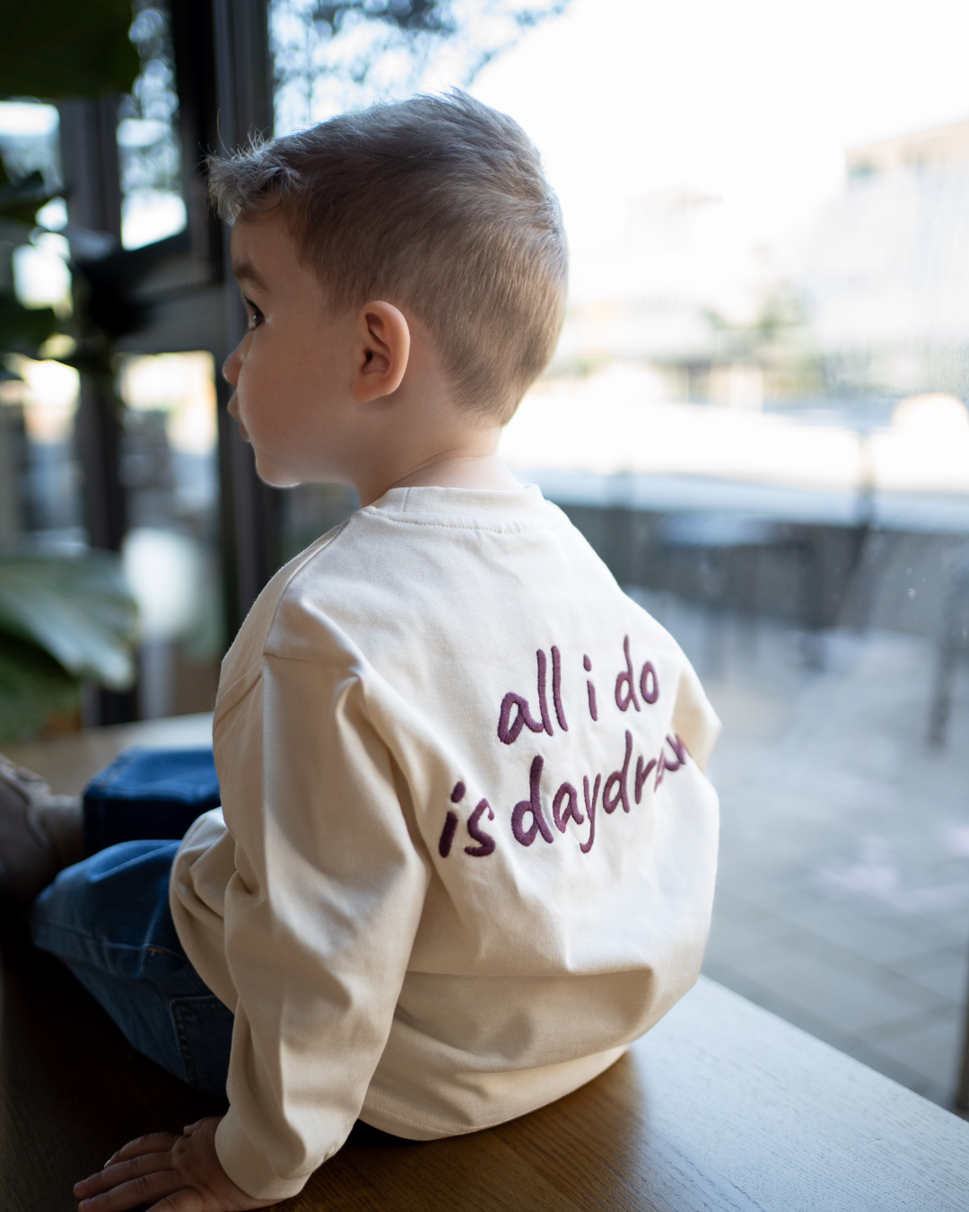 Junge sitzt seitlich am Fenster, cremefarbenes Daydream Longsleeve mit Rücken-Print „all i do is daydream" im warmen Gegenlicht.