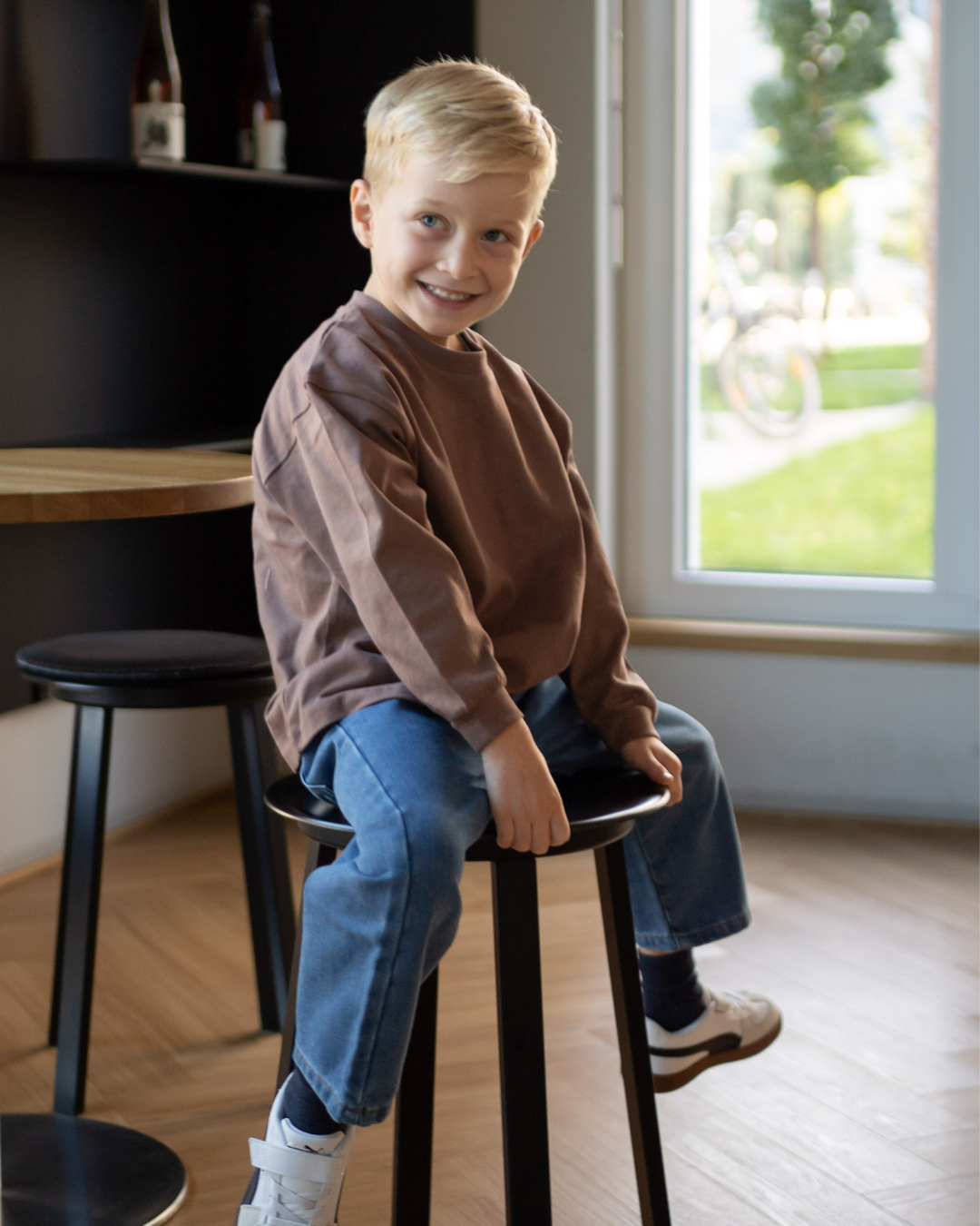 Lächelnder Junge sitzt auf einem Hocker im espressobraunen Comfy Studios Longsleeve und blauer Oversize-Jeans, Tageslicht am Fenster.