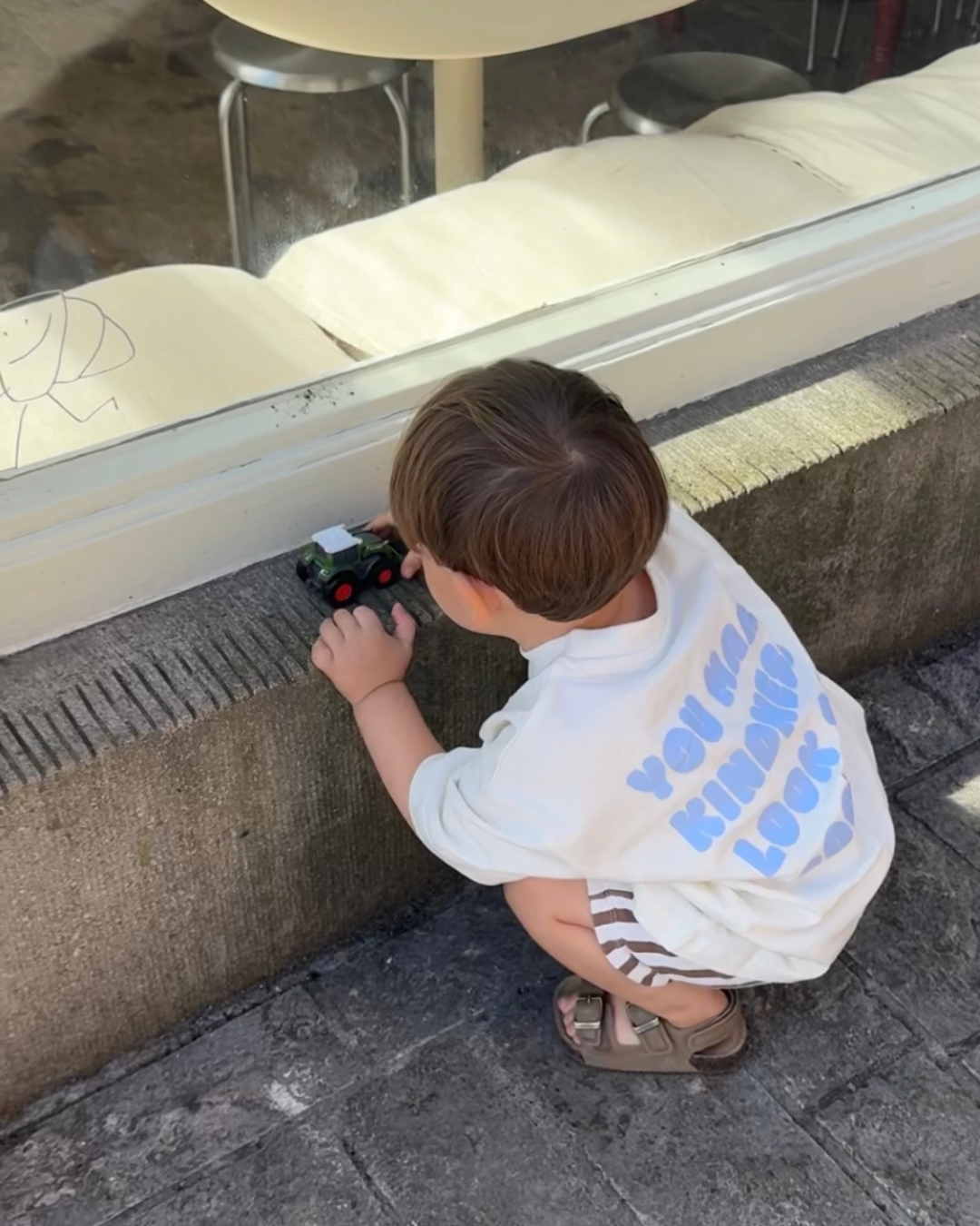 Kleinkind im oversize Kindness Shirt Blau von Comfy Studios mit blauem „You make kindness look so cool"-Print, spielt vor einem Café