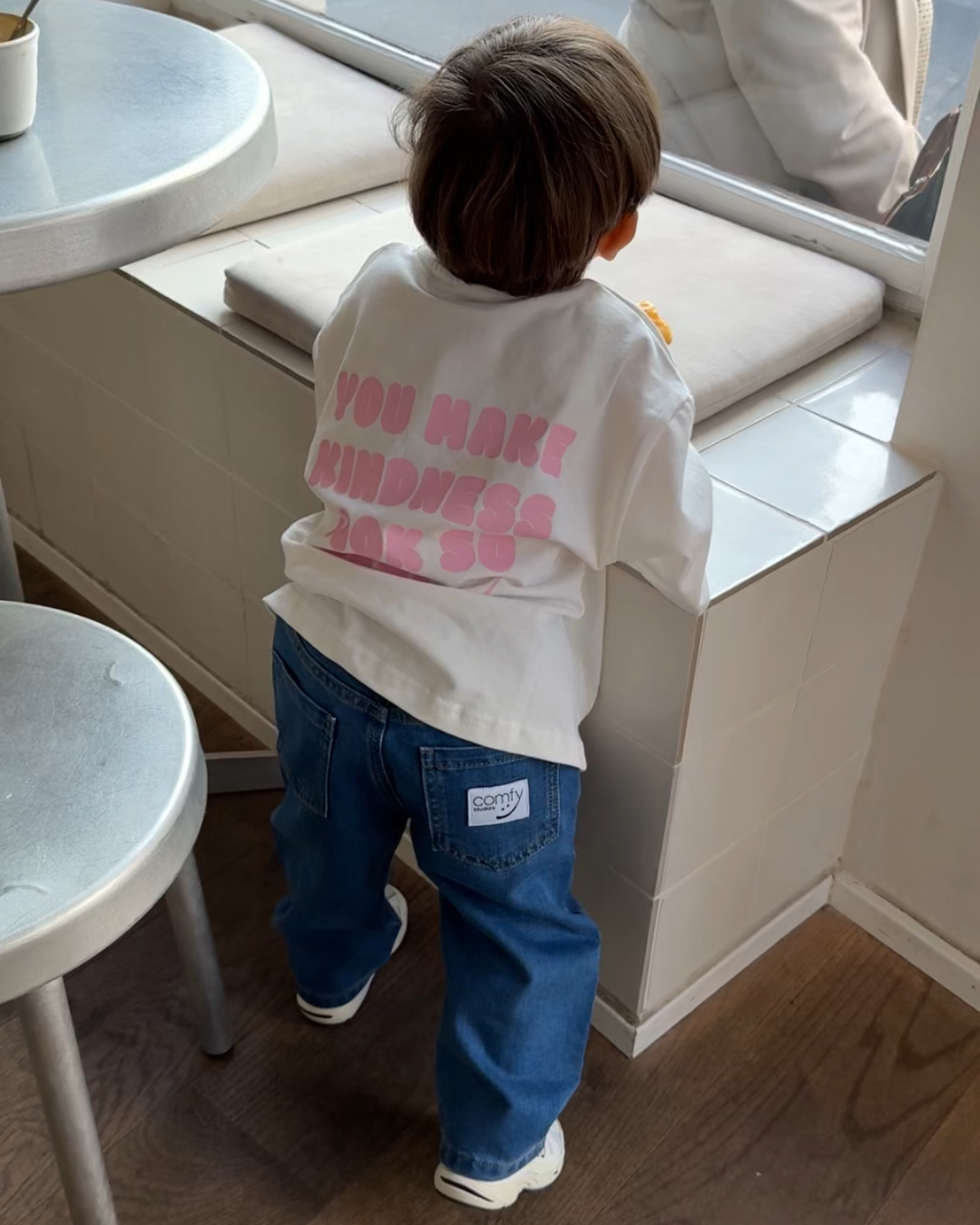 Kind klettert auf Bank im Café, trägt blaue Kinderjeans von Comfy Studios mit weißem Kindness Shirt