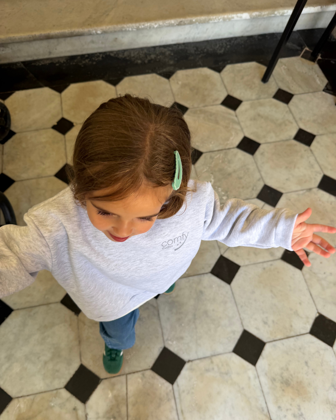 Mädchen in grauem Kinder-Sweatshirt von Comfy Studios streckt Arm aus, Comfy Studios Logo auf der Brust