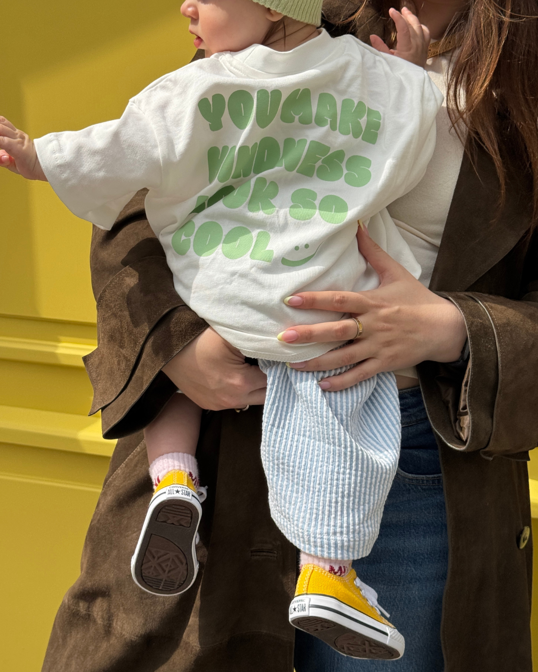 Kind im weißen Kindness Shirt mit grünem Rückenprint vor gelber Wand – Oversize-Kindershirt im Alltag.