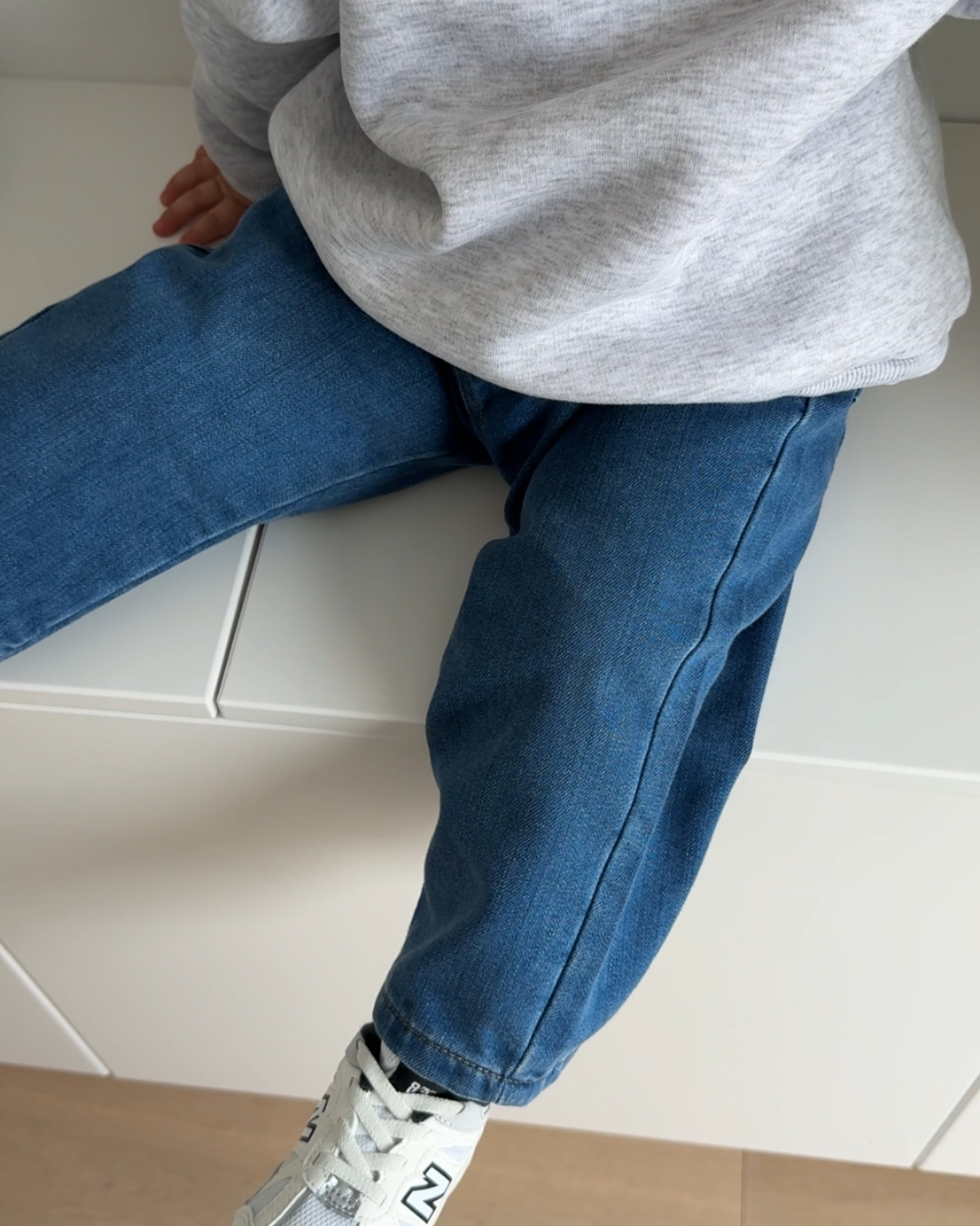 Kind sitzt lässig mit Comfy Studios Kindness Sweater und blauer Jeans, New Balance Sneakers