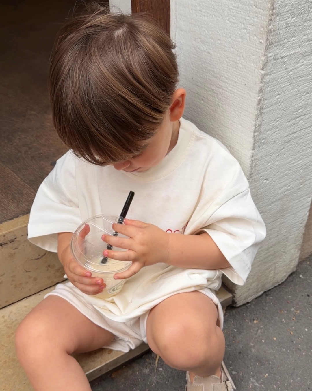 Kleines Kind sitzt auf Treppenstufen und trinkt aus Plastikbecher – weißes Oversize-Kindershirt von Comfy Studios, entspannter Sommermoment