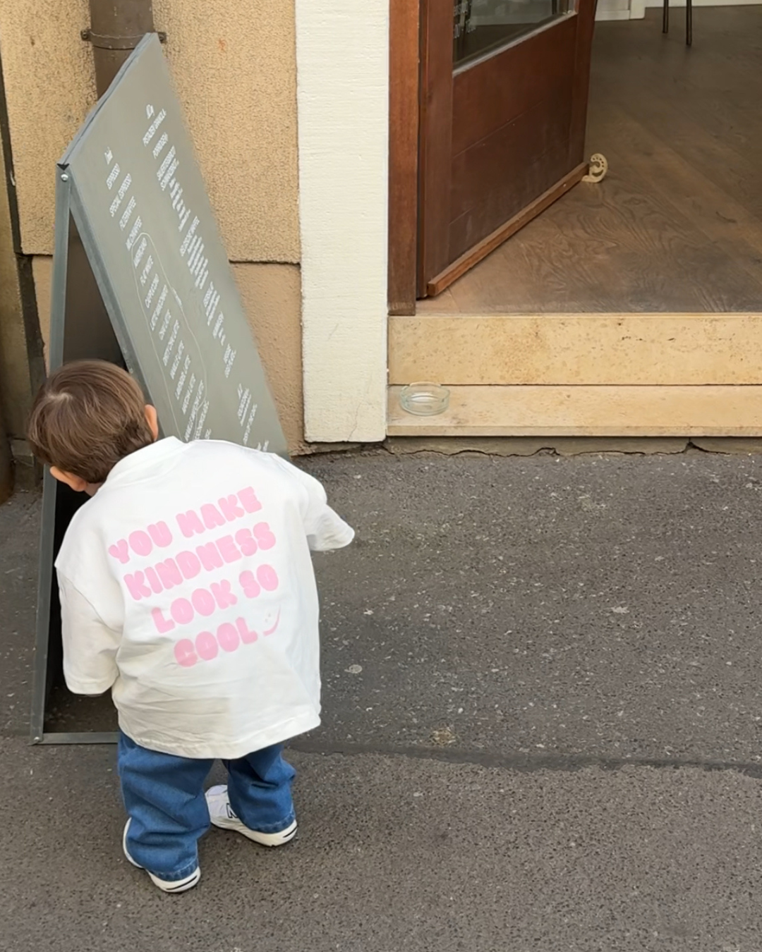 Kind von hinten im Kindness Shirt, neugierig an einer Café-Tafel – zeigt die Bewegungsfreiheit des oversize Schnitts.