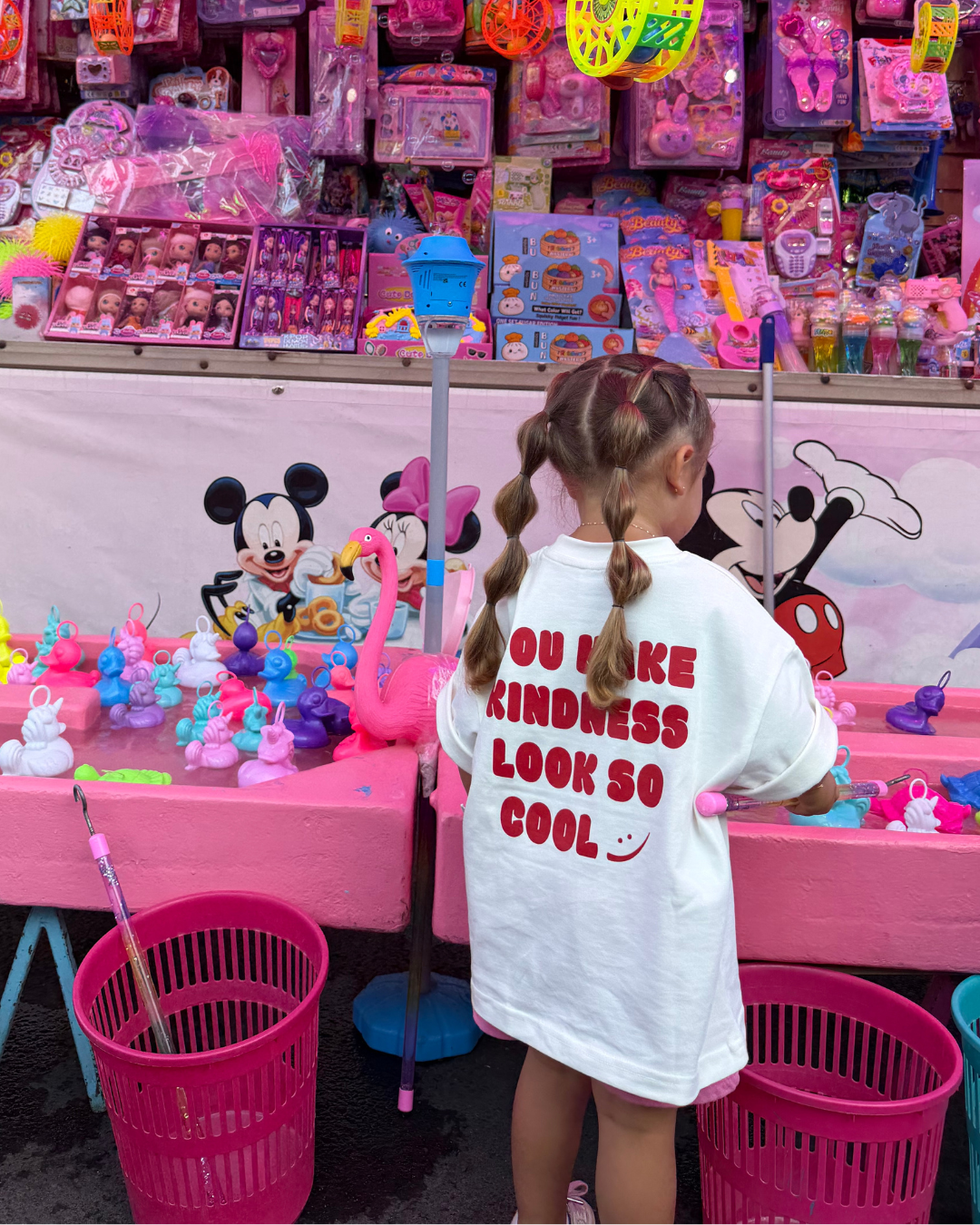 Kind steht vor buntem Jahrmarkt-Spielzeugstand, trägt weißes Kindness Shirt mit rotem Print You Make Kindness Look So Cool