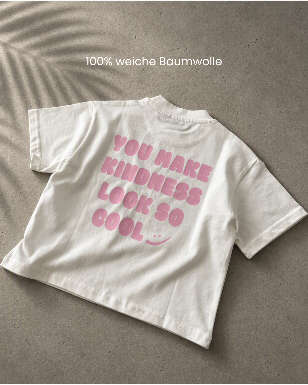 Weißes Kindness Shirt flach auf Betonoptik mit Hinweis ‚100 % weiche Baumwolle' und rosa Statement-Print auf der Rückseite.