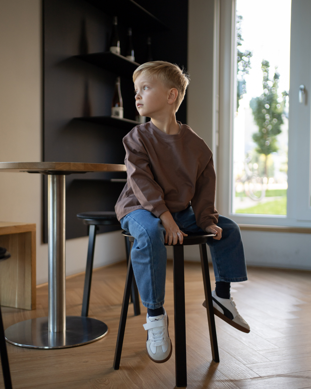 Junge sitzt auf einem Hocker am Fenster im kompletten Espresso-Look von Comfy Studios – espressobraunes Longsleeve und blaue Oversize-Jeans, warmes Tageslicht.