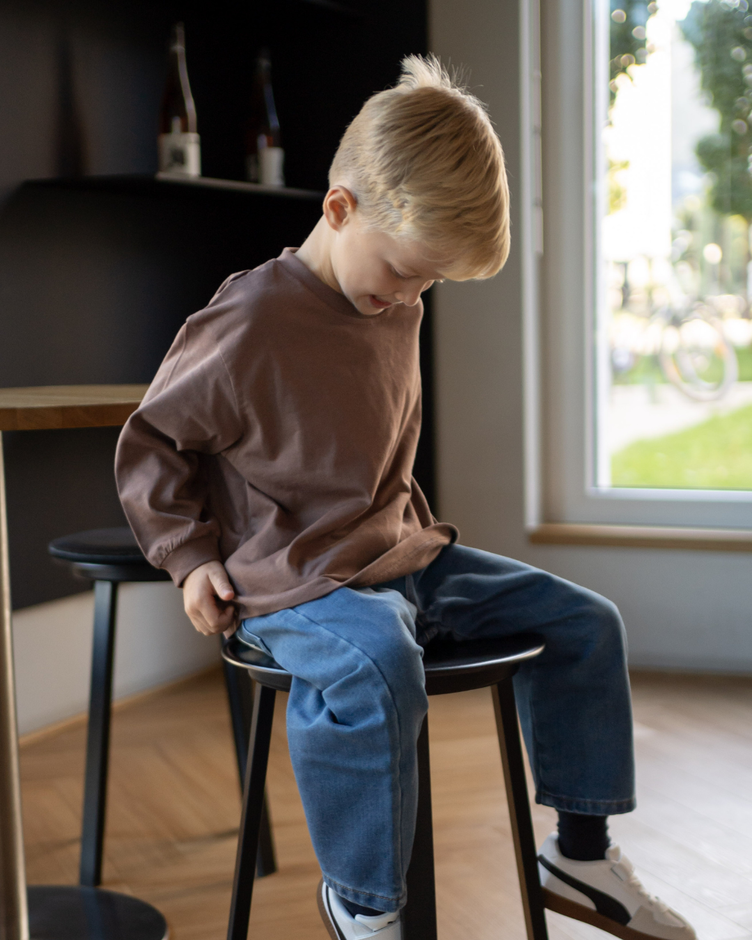 Junge sitzt auf einem Hocker im kompletten Espresso-Look von Comfy Studios, espressobraunes Longsleeve und blaue Oversize-Jeans.