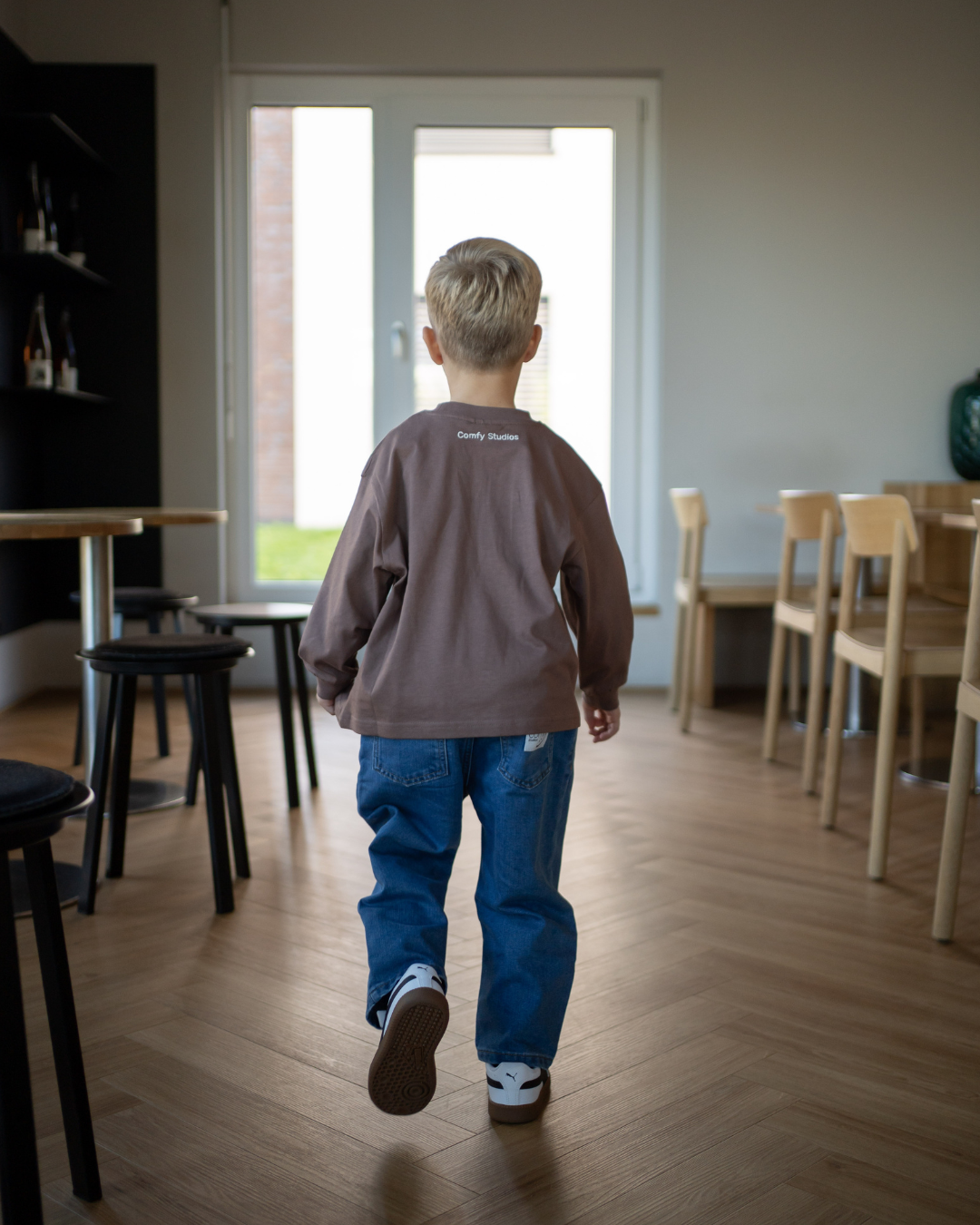 Junge von hinten im kompletten Espresso-Look, geht durch ein Café – espressobraunes Longsleeve mit „Comfy Studios"-Stickerei und blaue Jeans.