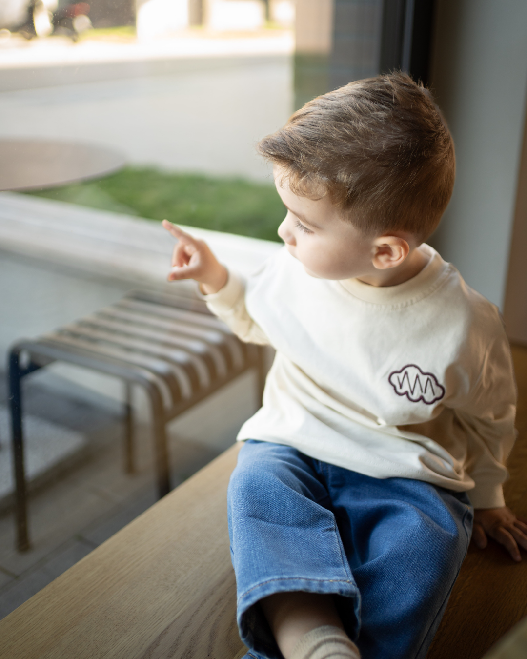 Kind sitzt am Fenster im cremefarbenen Daydream Longsleeve mit kleinem Brust-Logo und blauer Jeans – der komplette Look von Comfy Studios.