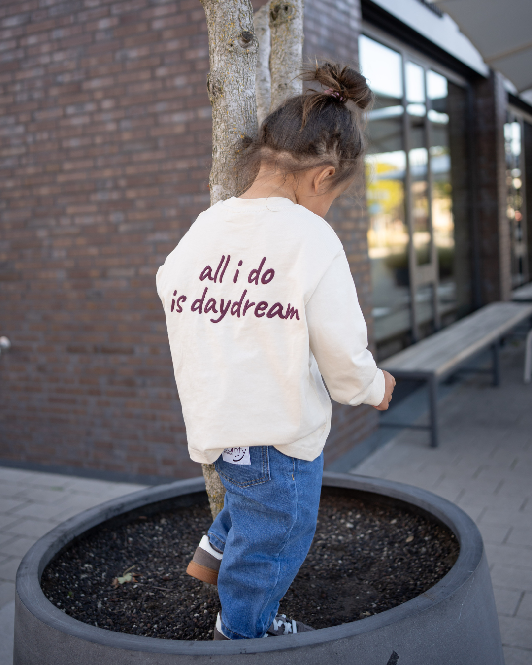 Kleinkind von hinten im cremefarbenen Daydream Longsleeve mit Rücken-Print „all i do is daydream", klettert draußen an einem Baum.