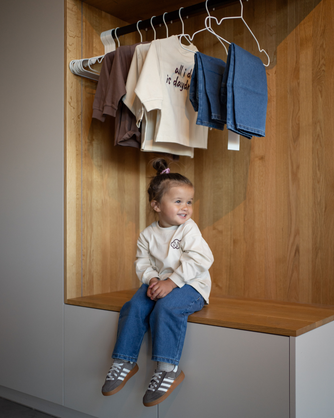Lächelndes Kleinkind sitzt im cremefarbenen Daydream Longsleeve und blauer Jeans, dahinter der Look am Kleiderbügel – Comfy Studios.