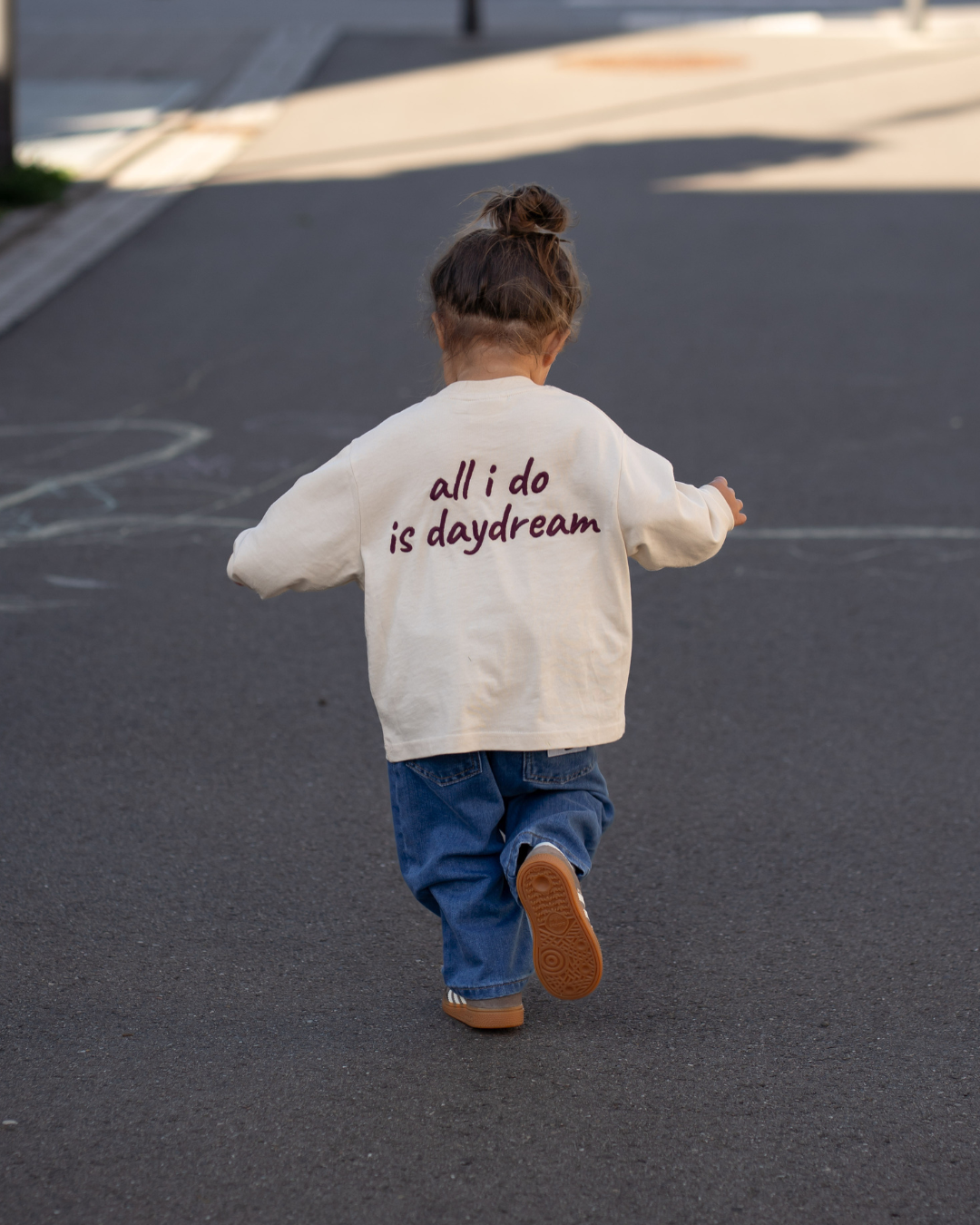 Kleinkind von hinten im cremefarbenen Daydream Longsleeve mit Rücken-Print „all i do is daydream", dazu blaue Oversize-Jeans, auf der Straße.