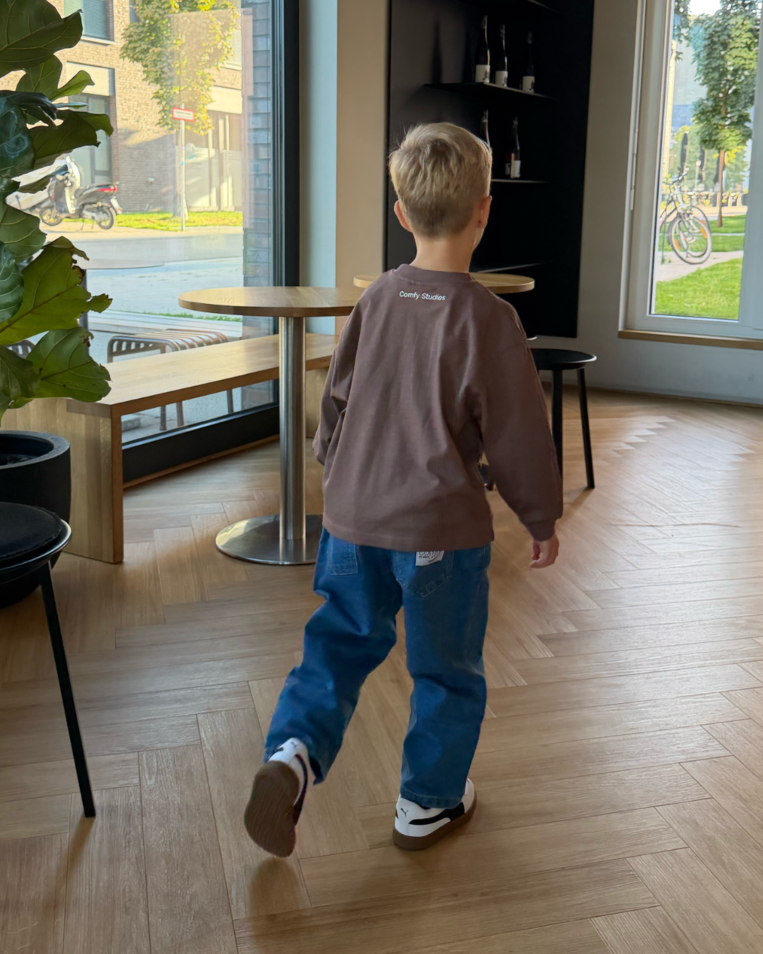 Kleinkind von hinten im espressobraunen Comfy Studios Longsleeve mit Marken-Stickerei am Rücken und blauer Jeans, in einem hellen Café.