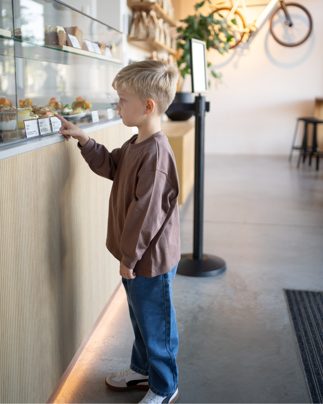 Junge steht seitlich an einer Café-Theke und zeigt auf Gebäck, im espressobraunen Comfy Studios Longsleeve mit blauer Oversize-Jeans.